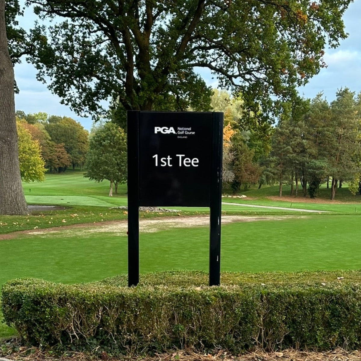 A black sign reading PGA  1st Tee stands on a landscaped entry area with trimmed hedges, a paved foreground, and a golf course with autumn trees in the background at the Belfry 
