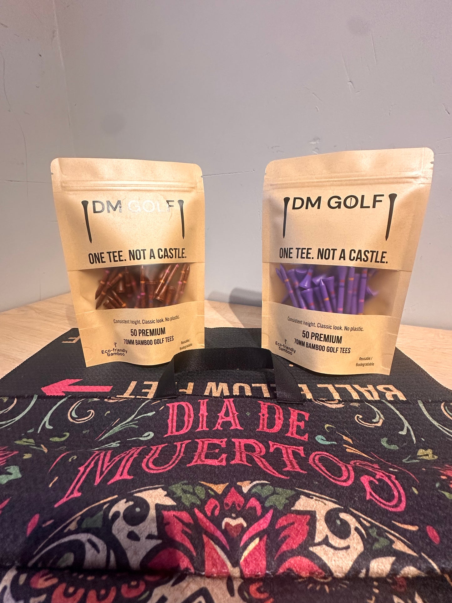 Two stand-up pouches of DM Golf Premium  Golf Ties are displayed on a black tote bag with a colorful Dia de Muertos design. The left pouch contains brown barbed golf ties, and the right pouch contains purple barbed golf ties.