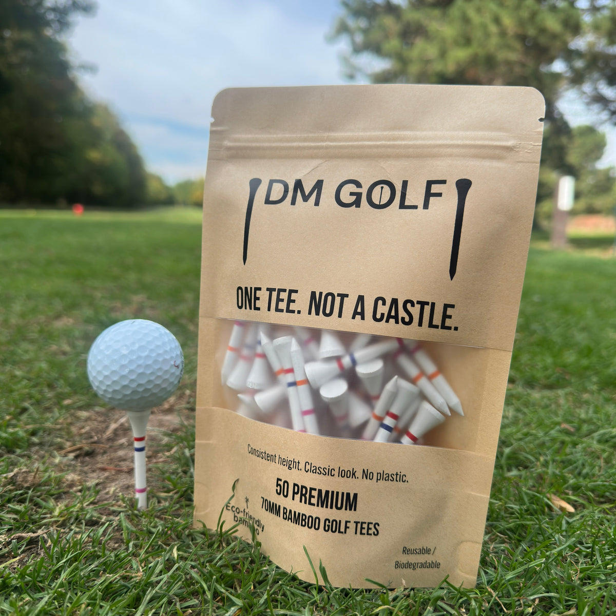 A stand-up pouch of DM Golf premium tees stands on grass at a golf course, next to a golf ball on a tee. The kraft paper bag has a clear window showing white tees inside and reads 'ONE TEE. A stand-up kraft pouch of DM Golf 7mm bamboo golf tees stands on a grassy golf course, next to a white golf ball on a tee. TheLink Not A Castle Tee, red, pink and orange castle tee height markers, one tee not 4, zero plastic, 
