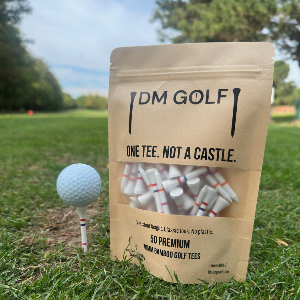 A stand-up pouch of DM Golf premium tees stands on grass at a golf course, next to a golf ball on a tee. The kraft paper bag has a clear window showing white tees inside and reads 'ONE TEE. A stand-up kraft pouch of DM Golf 7mm bamboo golf tees stands on a grassy golf course, next to a white golf ball on a tee. The Link Not A Castle Tee, red, pink and orange castle tee height markers, one tee not 4, zero plastic,