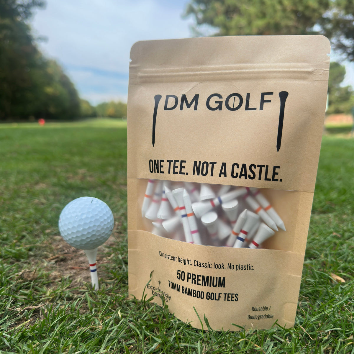 A stand-up pouch of DM Golf premium tees stands on grass at a golf course, next to a golf ball on a tee. The kraft paper bag has a clear window showing white tees inside and reads 'ONE TEE. A stand-up kraft pouch of DM Golf 7mm bamboo golf tees stands on a grassy golf course, next to a white golf ball on a tee. The Link Not A Castle Tee, red, pink and orange castle tee height markers, one tee not 4, zero plastic, Set to pink castle tee 