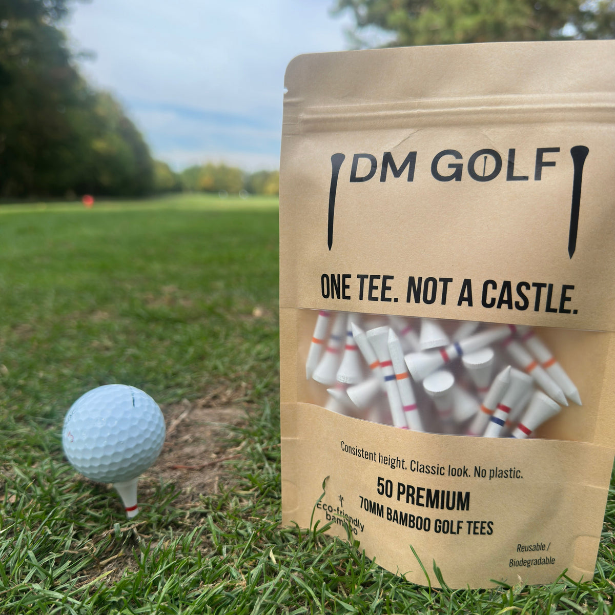 A stand-up pouch of DM Golf premium tees stands on grass at a golf course, next to a golf ball on a tee. The kraft paper bag has a clear window showing white tees inside and reads 'ONE TEE. A stand-up kraft pouch of DM Golf 7mm bamboo golf tees stands on a grassy golf course, next to a white golf ball on a tee. The Link Not A Castle Tee, red, pink and orange castle tee height markers, one tee not 4, zero plastic, set to blue castle tee