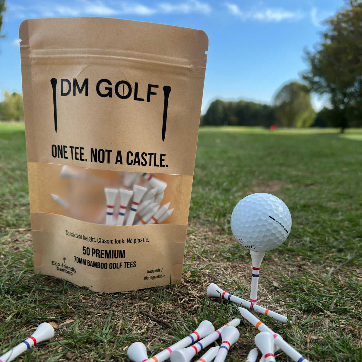 A stand-up pouch of DM Golf premium tees stands on grass at a golf course, next to a golf ball on a tee. The kraft paper bag has a clear window showing white tees inside and reads 'ONE TEE. A stand-up kraft pouch of DM Golf 7mm bamboo golf tees stands on a grassy golf course, next to a white golf ball on a tee. The Link Not A Castle Tee, red, pink and orange castle tee height markers, one tee not 4, zero plastic,