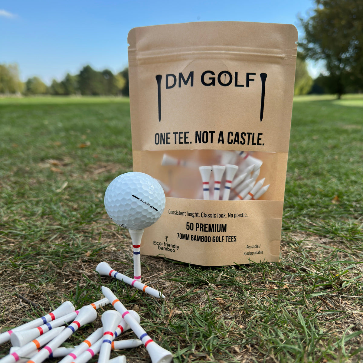 A stand-up pouch of DM Golf premium tees stands on grass at a golf course, next to a golf ball on a tee. The kraft paper bag has a clear window showing white tees inside and reads 'ONE TEE. A stand-up kraft pouch of DM Golf 7mm bamboo golf tees stands on a grassy golf course, next to a white golf ball on a tee. The Link Not A Castle Tee, red, pink and orange castle tee height markers, one tee not 4, zero plastic, pink castle tee 